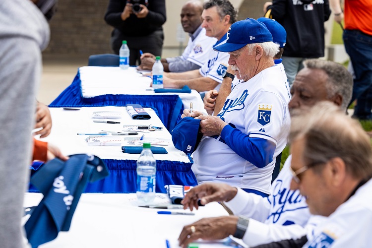 Six former Royals players sit in a line at tables to sign autographs. All are wearing Royals jerseys.