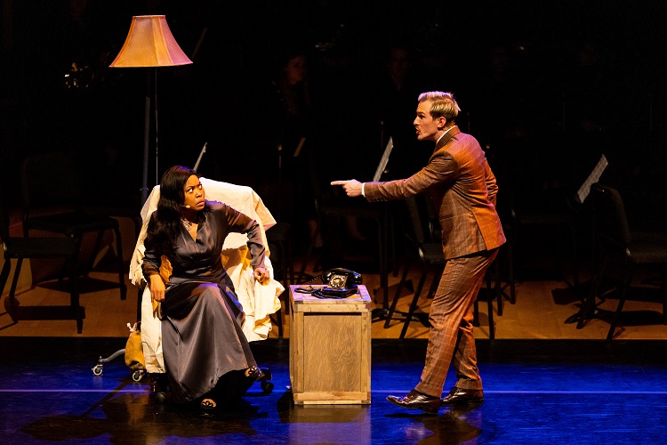 Two students act out a scene on stage - on is sitting in an armchair and one is standing next to it and pointing at her. A side table with a lamp is beside the arm chair.