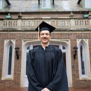 Cody Truitt stands in front of a building on UMKC Volker campus. He looks proud and wears a graduation cap and gown.