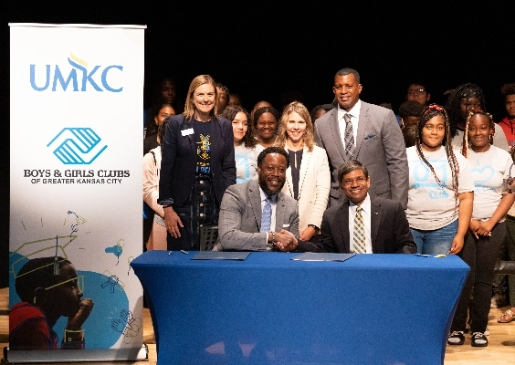 The UMKC Chancellor and representative from Boys and Girls Clubs of Kansas City shake hands a table with other UMKC representatives and a youth group behind them.