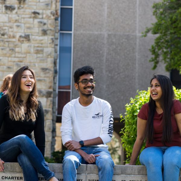 Three international students sit on a rock wall on UMKC Volker campus. They are laughing and enjoying a sunny fall day.
