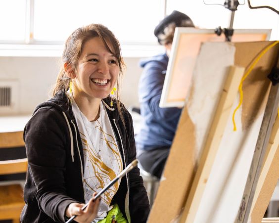 student looking happy sits at an easel with a paintbrush in hand