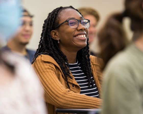 a student smiles broadly as she sits in class surrounded by other students. She wears glasses and a cardigan.