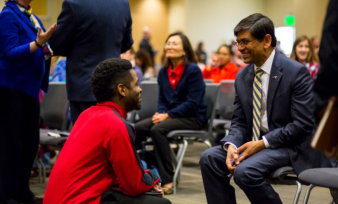 Student presenting as black male squats in front of seated and smiling Chancellor Agrawal