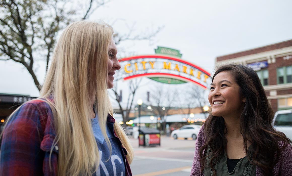 Two students presenting as women stand in front of City Market sign