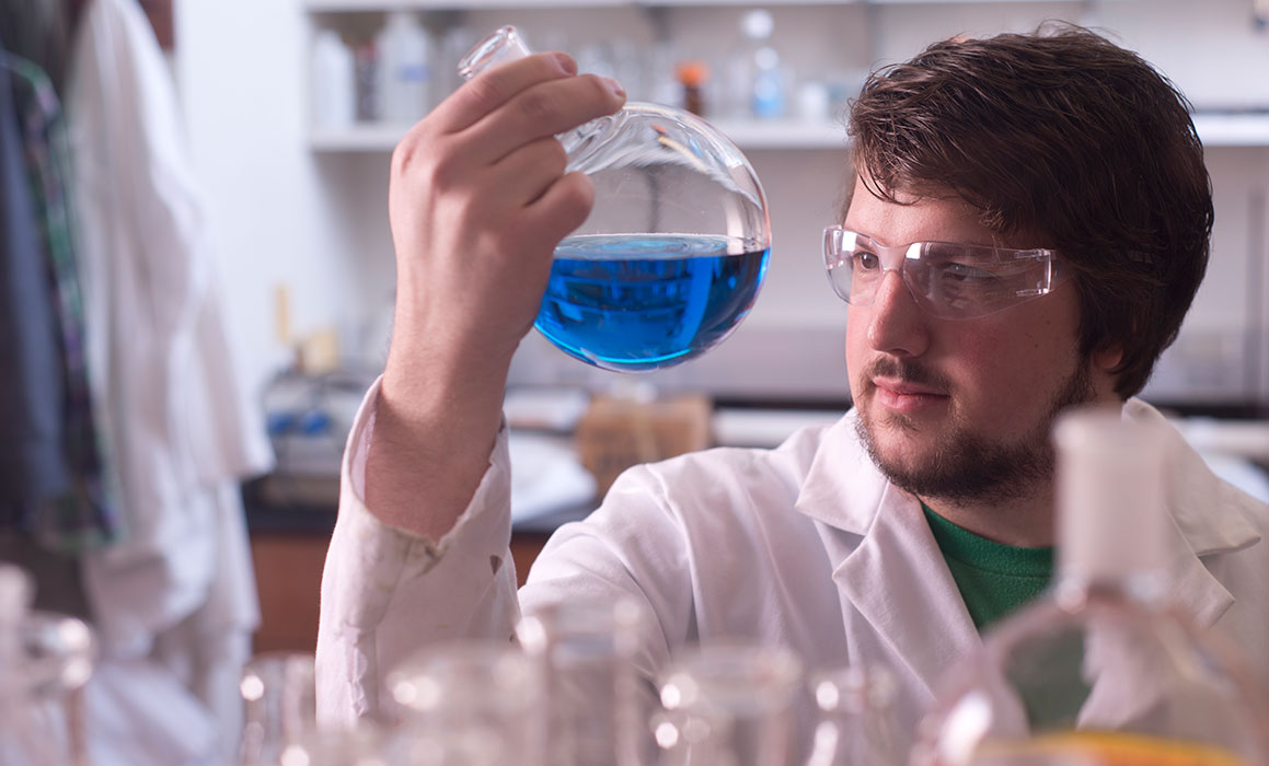 Student presenting as white male in lab coat and safety glasses holds up and looks at round vial with blue liquid in a lab