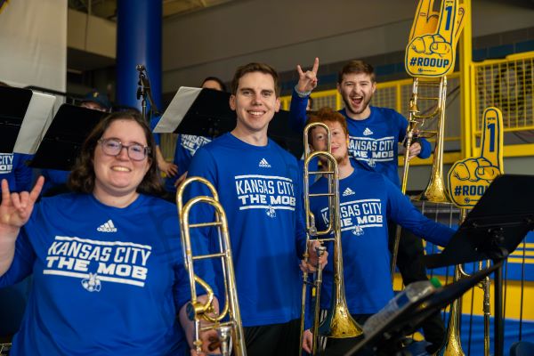 Members of the Mob, the UMKC pep band, wear blue shirts and make the Roo Up hand sign in the stands for a basketball game at Swinney.