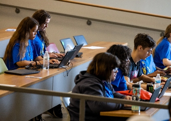 students sit in a lecture hall on risers with laptops open. Many have water bottles.