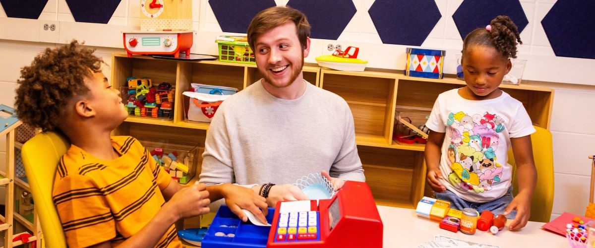 An education student interacts with two children in the play therapy room. They have a toy cash register in front of them on a table and look like they're having fun.