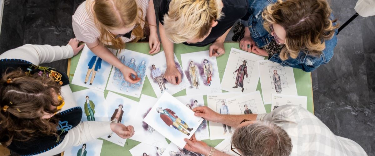 An overhead view of sketches on a table surrounded by students and the professor in a costume shop session