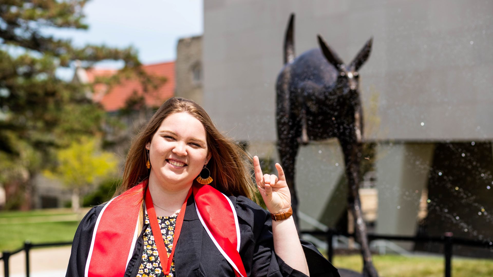 student wearing graduation gown makes the roo up gesture with her hand while posing in front of the Corbin roo statue