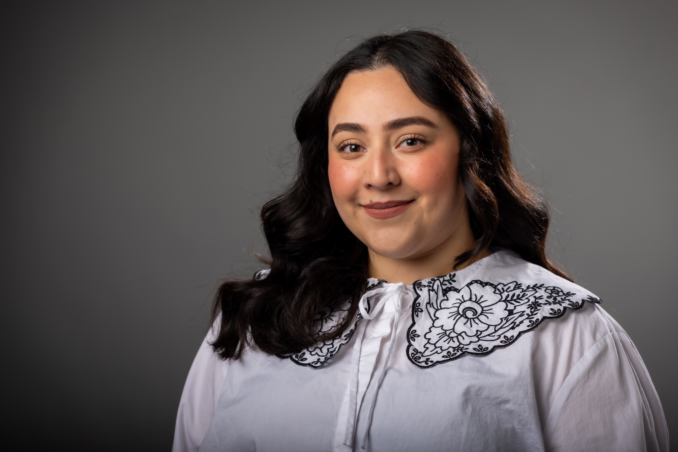 Victoria Dominguez studio headshot, young person with long dark hair smiling at the camera in front of a gray portrait backdrop