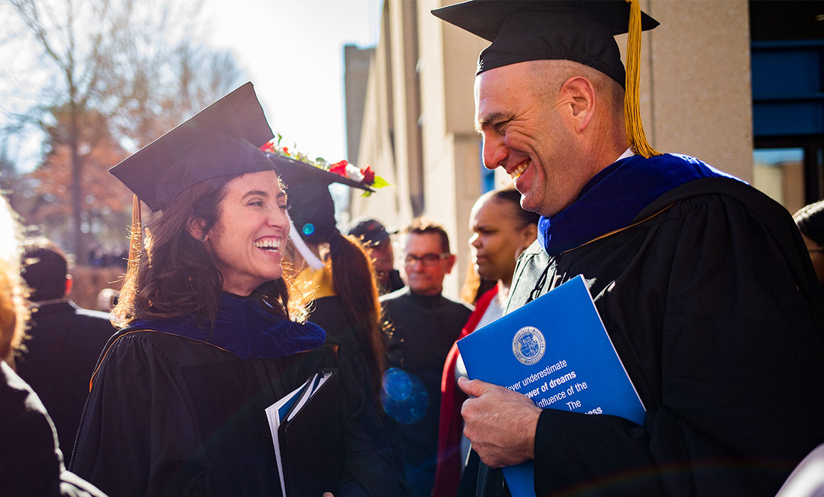 Person presenting as white woman in graduation cap and gown smiles at person presenting as white man wearing cap and gown