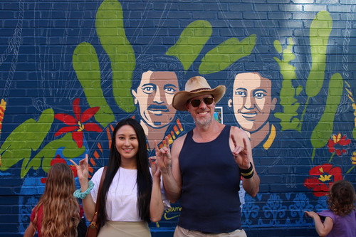 Two people stand in front of a mural-in-progress on a brick wall. They are making the Roo Up hand gesture.