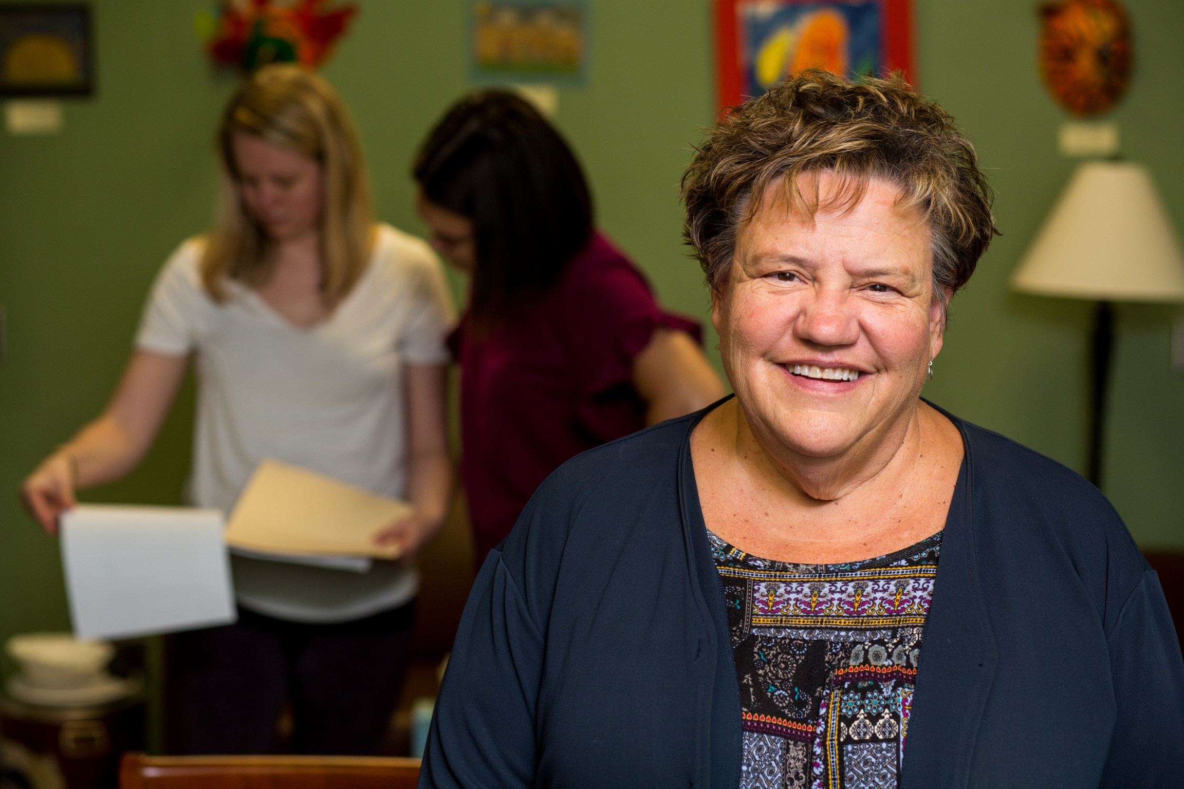 Mary Kay O'Malley smiles while sitting in the Child and Family Services Clinic in the law school. Two students are in the background, looking at paperwork.