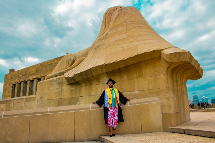 Nursing Alumnus Coco Ndipagbor stands in her graduation gown in front of one of the Assyrian Sphinxes at the Liberty Memorial