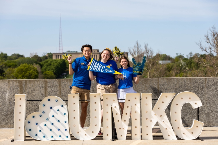 two students wearing blue shirts stand behind a light up sign that reads 'I <3 UMKC'