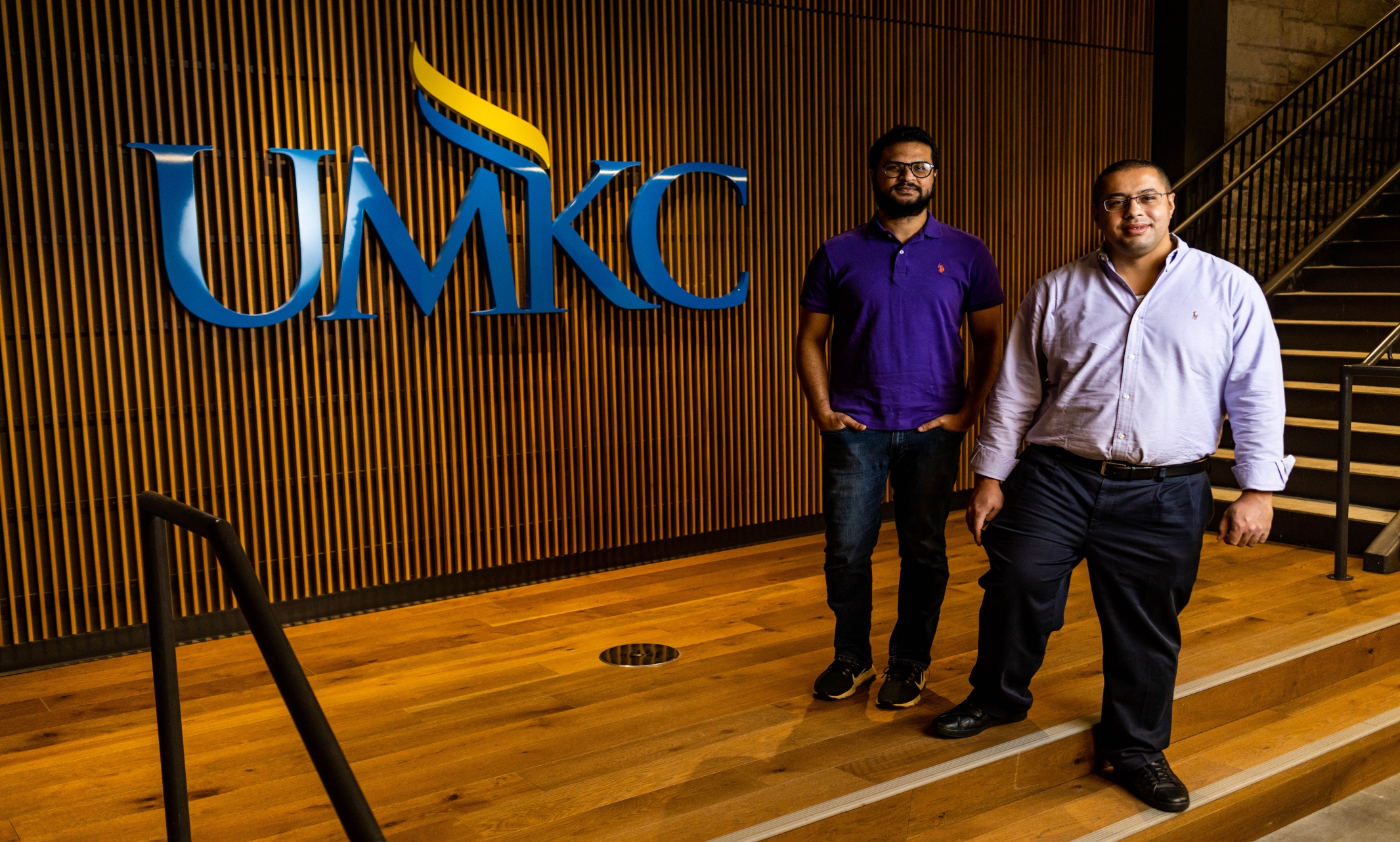 Two men stand in front of a wood wall with the letter 'UMKC' in blue behind them