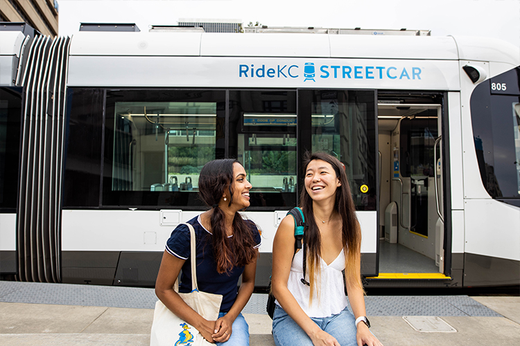 Two smiling students sit in front of the KC Streetcar