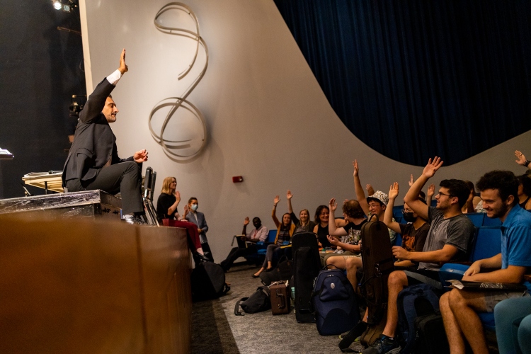 A conservatory professor sits on a stage with his feet dangling addressing students sitting in an auditorium. Nearly all of them have their hand raised. 