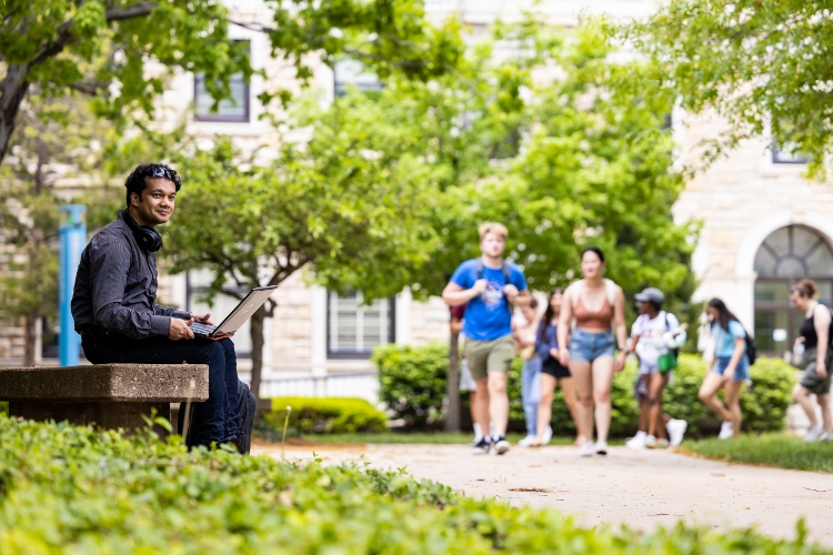 A student sits outside on a bench with a laptop on his lap. A group of students on the walkway behind him is walking his direction