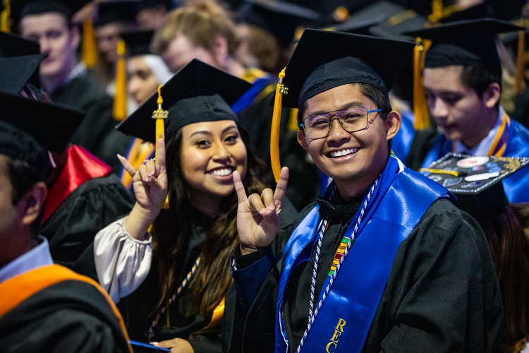 Two graduates do a Roo Up hand gesture in their regalia