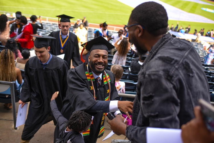 A graduate walks up the stairs after receiving his diploma and shakes hands with a loved one as a young boy hugs him