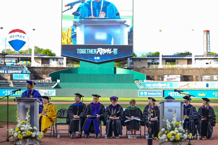 Chancellor Agrawal stands at the podium with the deans seated behind him on the field of Kauffman Stadium
