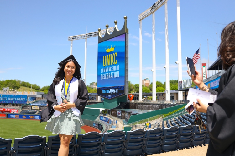 A graduate stands in front of Crown Vision at Kauffman Stadium as someone takes her photograph