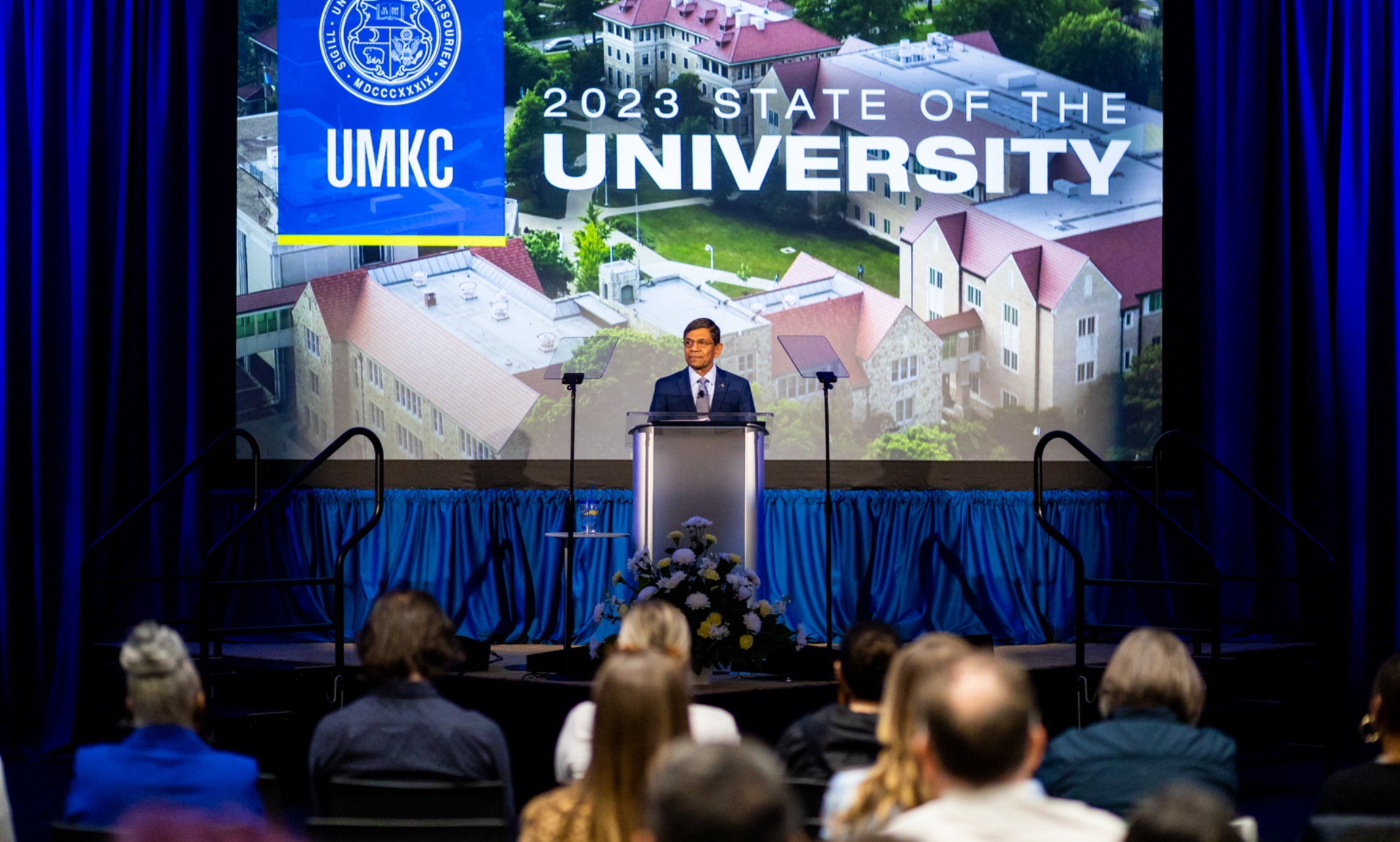 Chancellor C. Mauli Agrawal stands in front of a crowd. Behind him an aerial view of campus is displayed on a screen with "State of the University and the UMKC Seal imposed on the foreground