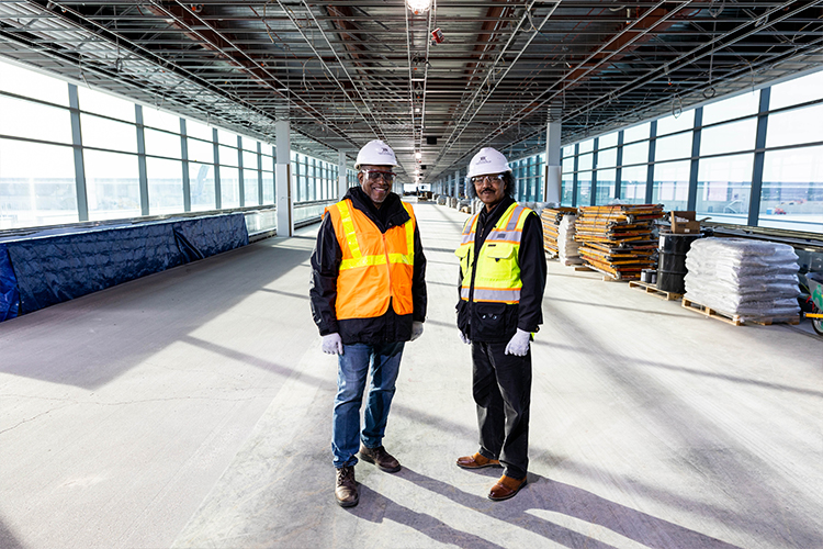 Leonard Graham and Hagos Andebrhan stand together in a large space under construction, wearing construction gear.