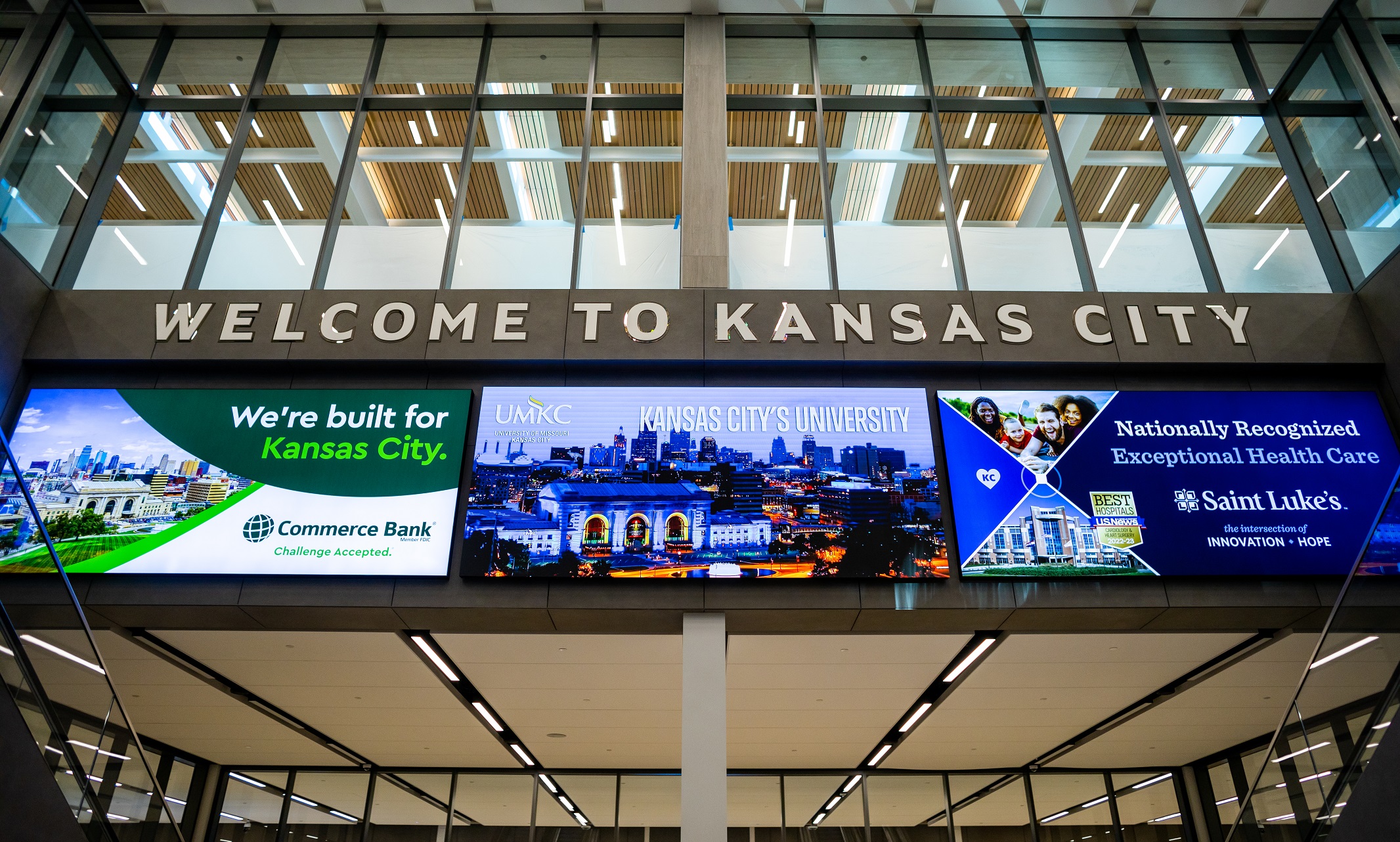 A sign over escalators down to baggage claim reads "Welcome to Kansas City." Below it are three large video boards for advertising. The middle board reads, "UMKC, Kansas City's University."