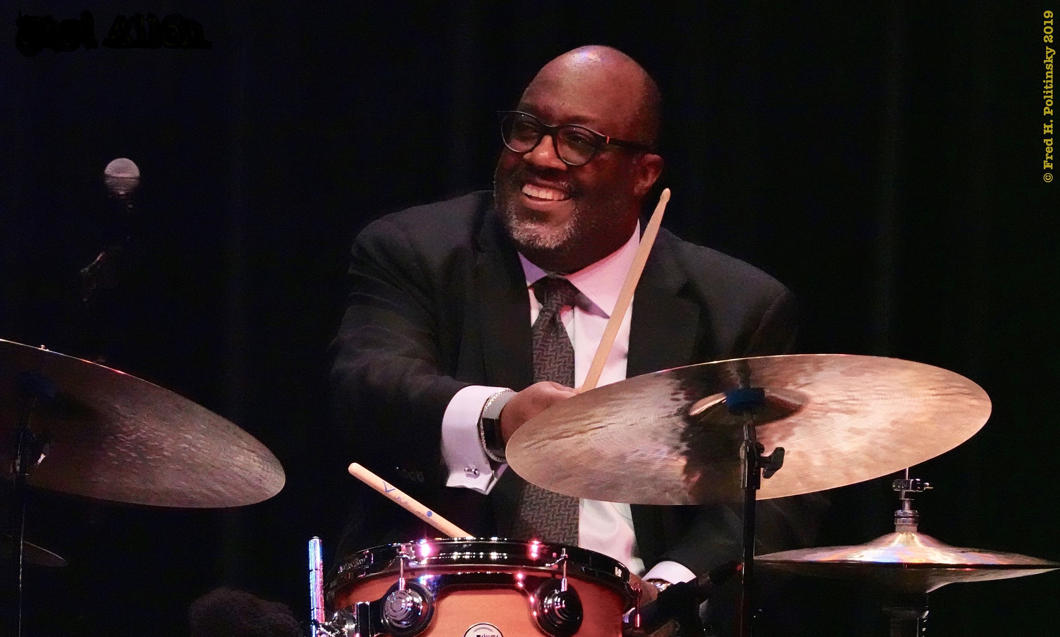 Carl Allen sits behind a drum set with drum sticks in his hand and smiles while looking to the side and playing