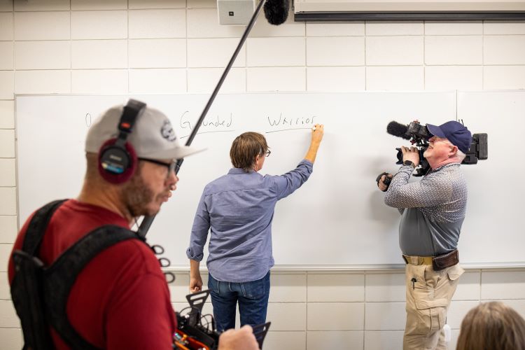 Clancy Martin writes on the whiteboard while teaching a class; CBS cameras and reporters are around him