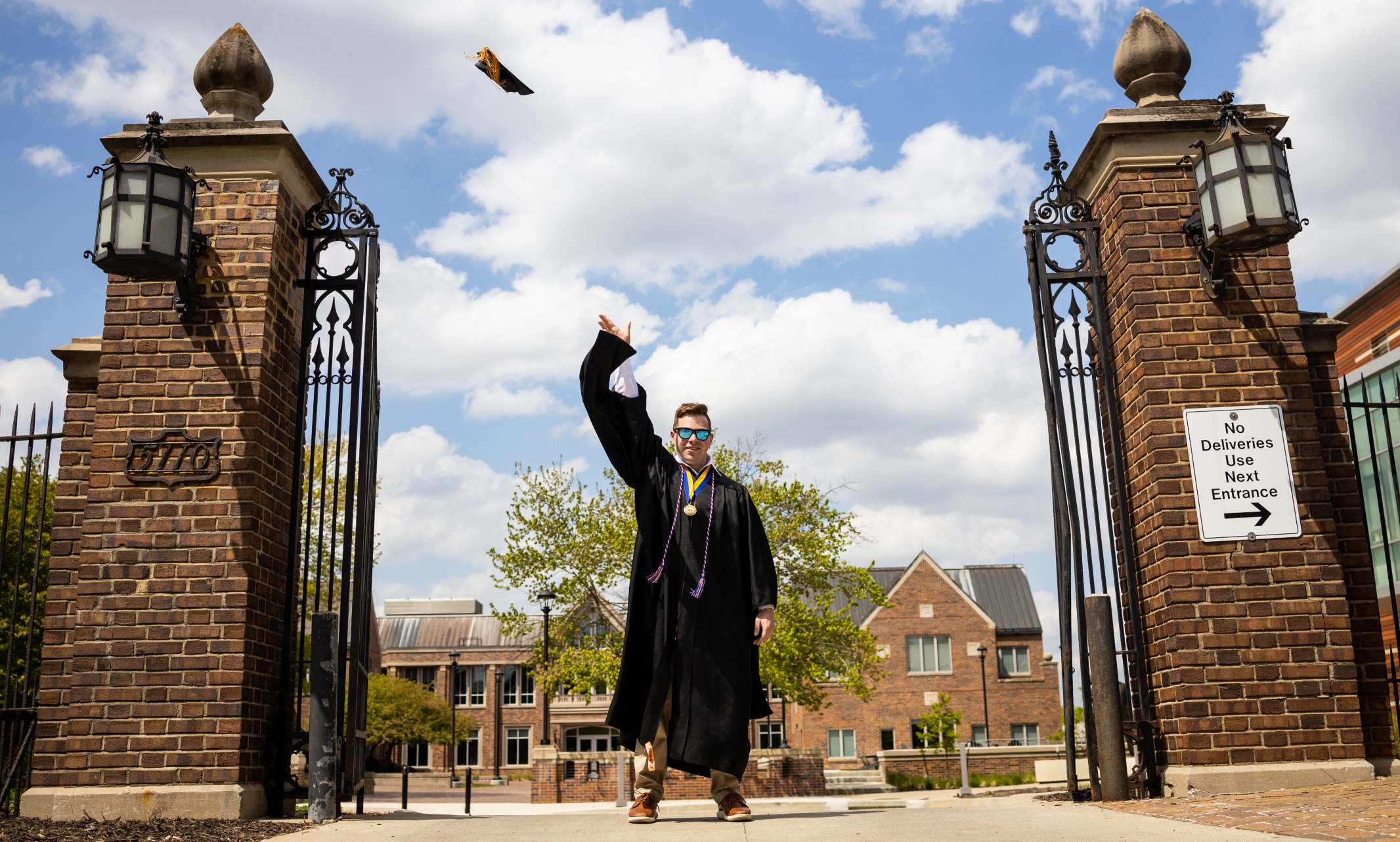 Henry Meeds stands at the center of the gate in front of Bloch Heritage Hall. He is dressed in commencement regalia and wearing sunglasses, throwing the graduation cap in the air.