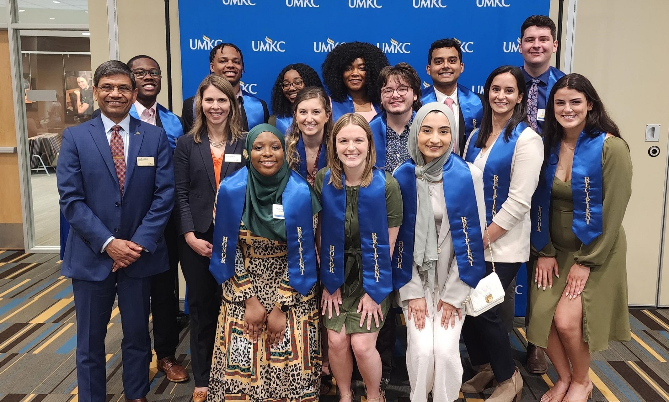Thirteen students stand with Chancellor Agrawal and Provost Lundgren in front of a UMKC backdrop and smile