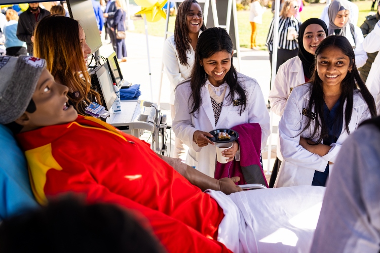 Several students in white coats laugh, standing behind a mannequin that is dressed in Chiefs gear and laying on a hospital bed
