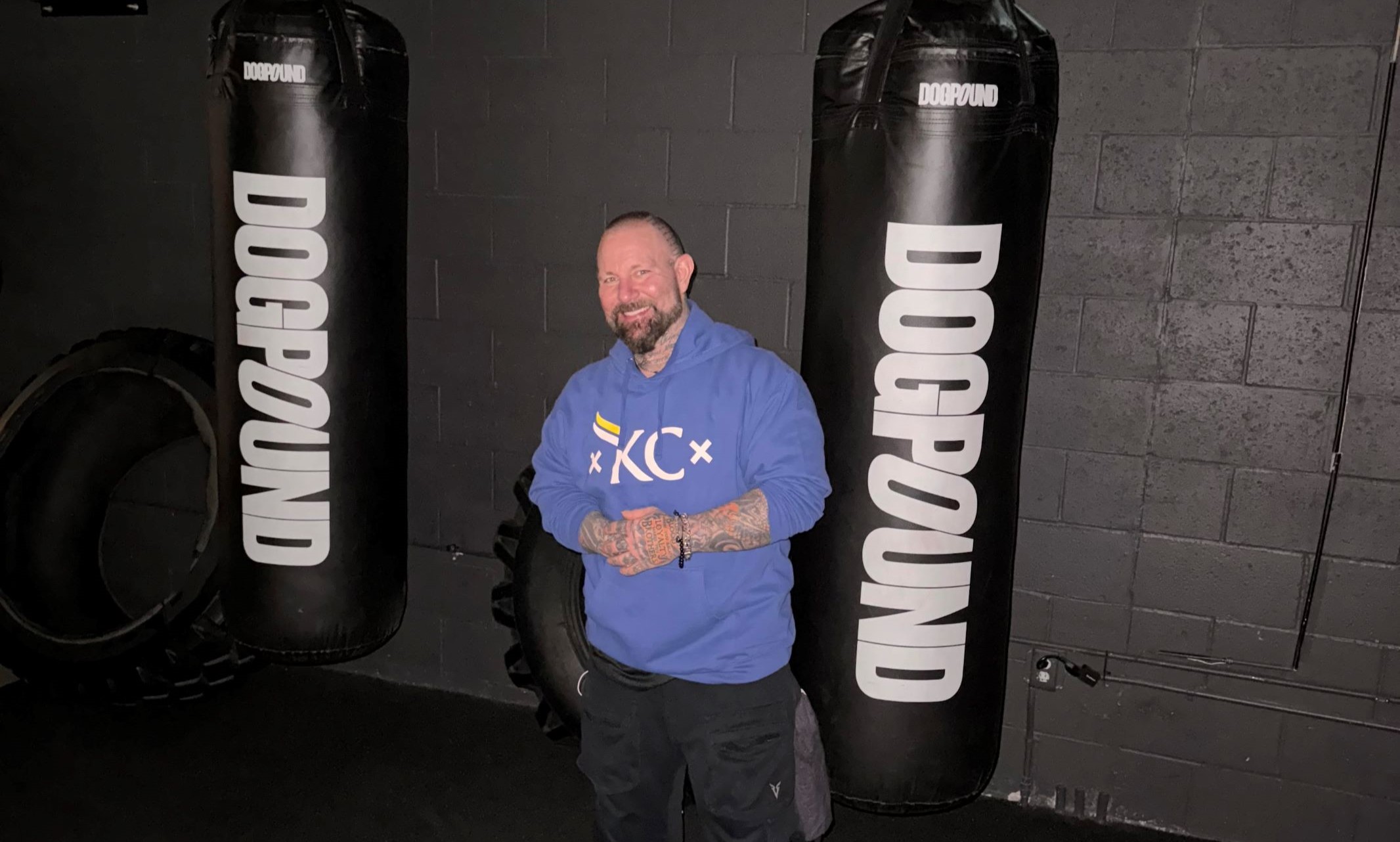 Taylor Swift and other celebrities' fitness trainer UMKC Alum Kirk Myers wearing a blue KC Mobb Made Sweatshirt in front of boxing sandbags at his gym dogpound