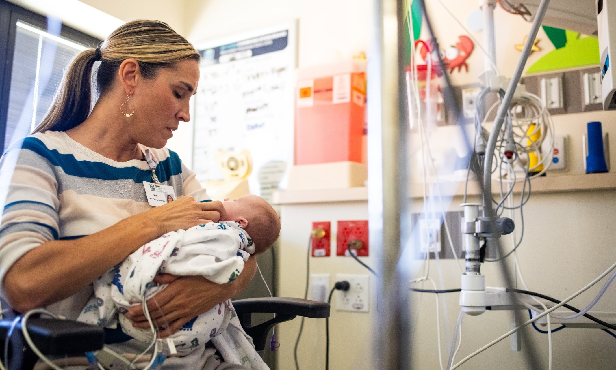 Amy Robertson holds a very small baby while sitting in a hospital room. The baby's NICU crib and monitors are visible beside them