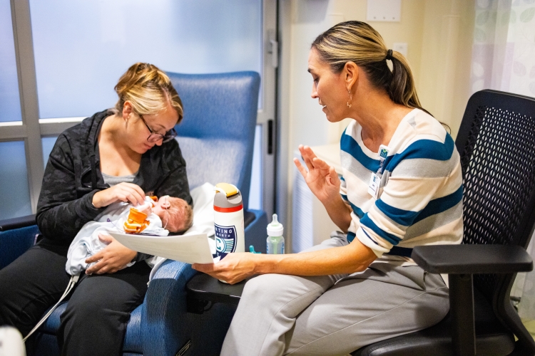 Amy Robertson holds a paper out for a mother to see and talks as the mother holds her baby in the NICU