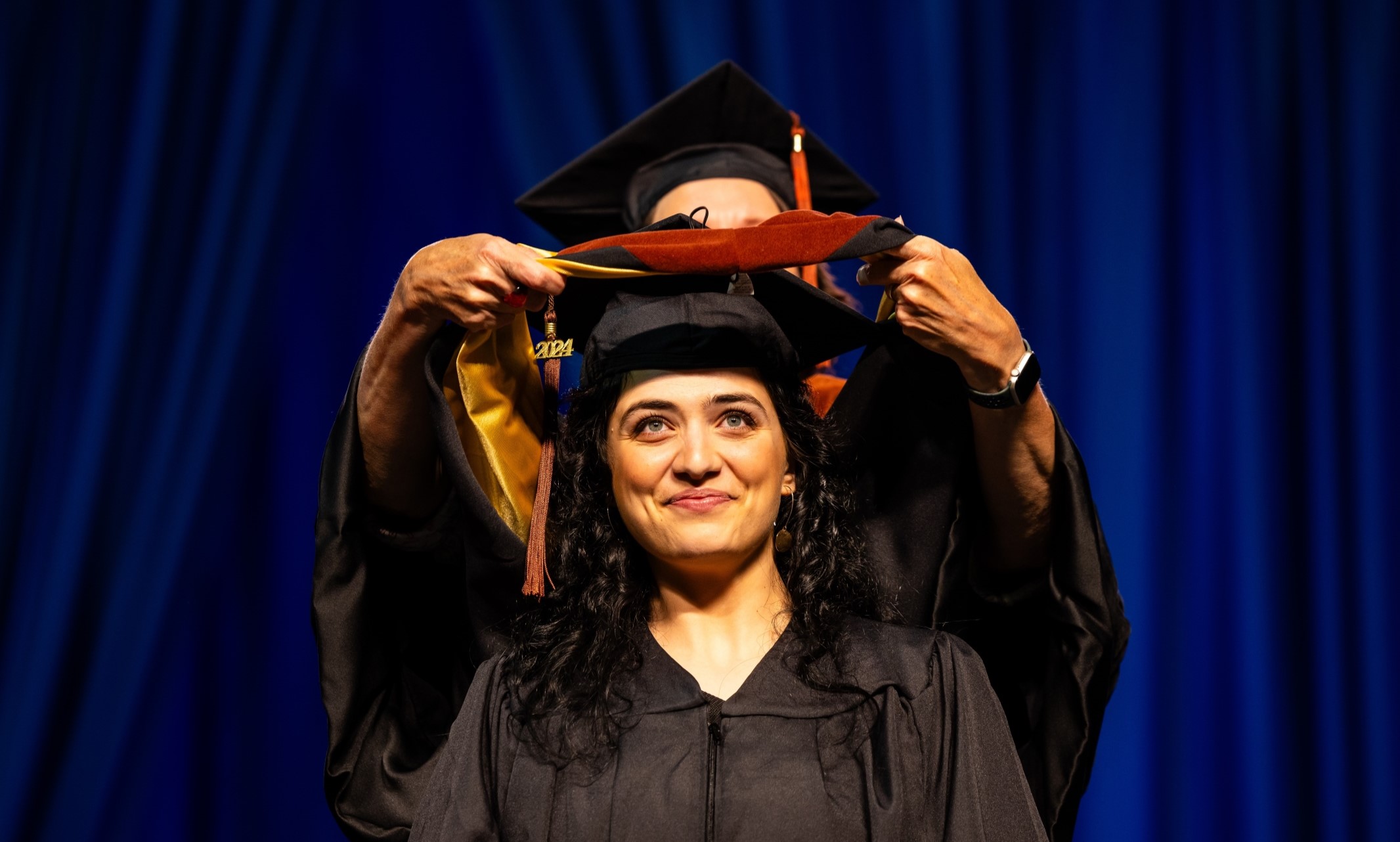 A Ph.D. student stands on stage at Commencement, a professor stands behind her and lowers the doctoral hood over the student's head in the hooding ceremony