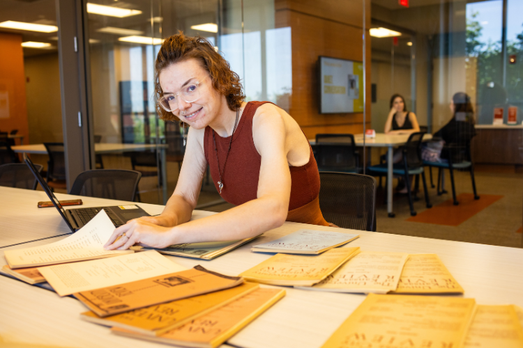 Student with documents on the table and a laptop open. She is smiling at the camera.