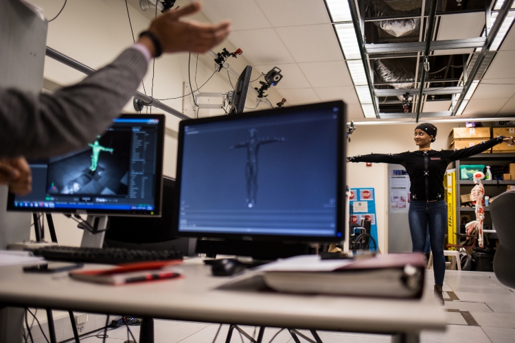 A student is hooked up to a motion capture machine with arms outstretched. A computer screen in the foreground shows a digital model that matches the student's stance.