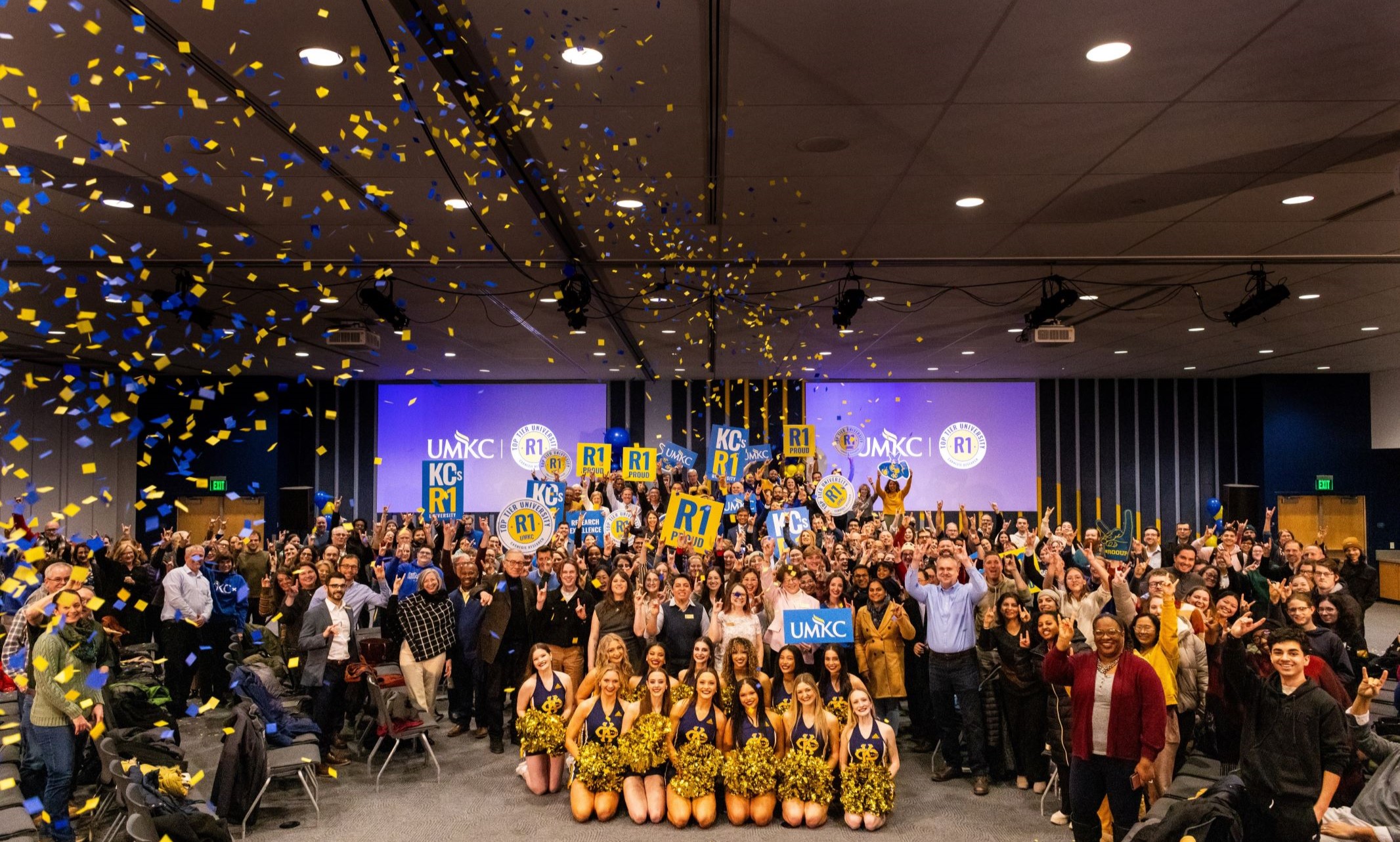 A large crowd of several hundred people gather together for a photo. People in the middle hold up "R1 Proud" and UMKC signs and many people around the room hold their hands in a Roo Up gesutre. Blue and yellow confetti falls down on them from the left side of the photo.