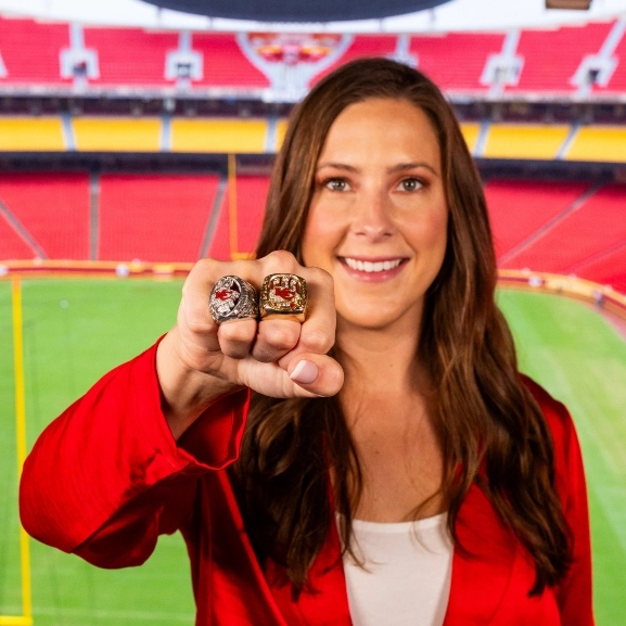 Meghan Jaben showing off her Super Bowl rings in front of GEHA field at Arrowhead stadium.